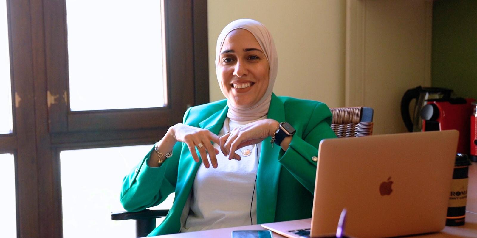 Woman in green jacket and white hijab sits at a desk smiling