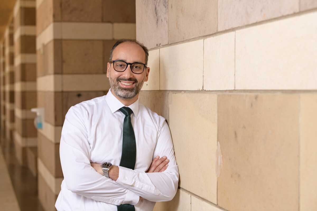 Smiling man in glasses wearing a button-up and blue tie leaning against a wall at AUC