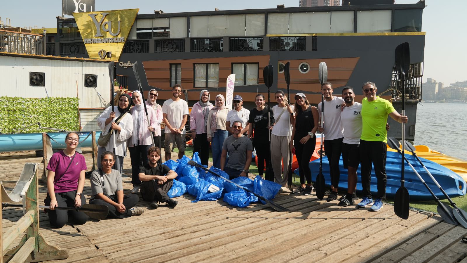 A group of students and faculty standing together on Qursaya Island with plastic bags next to them that have trash colelcted from the Nile