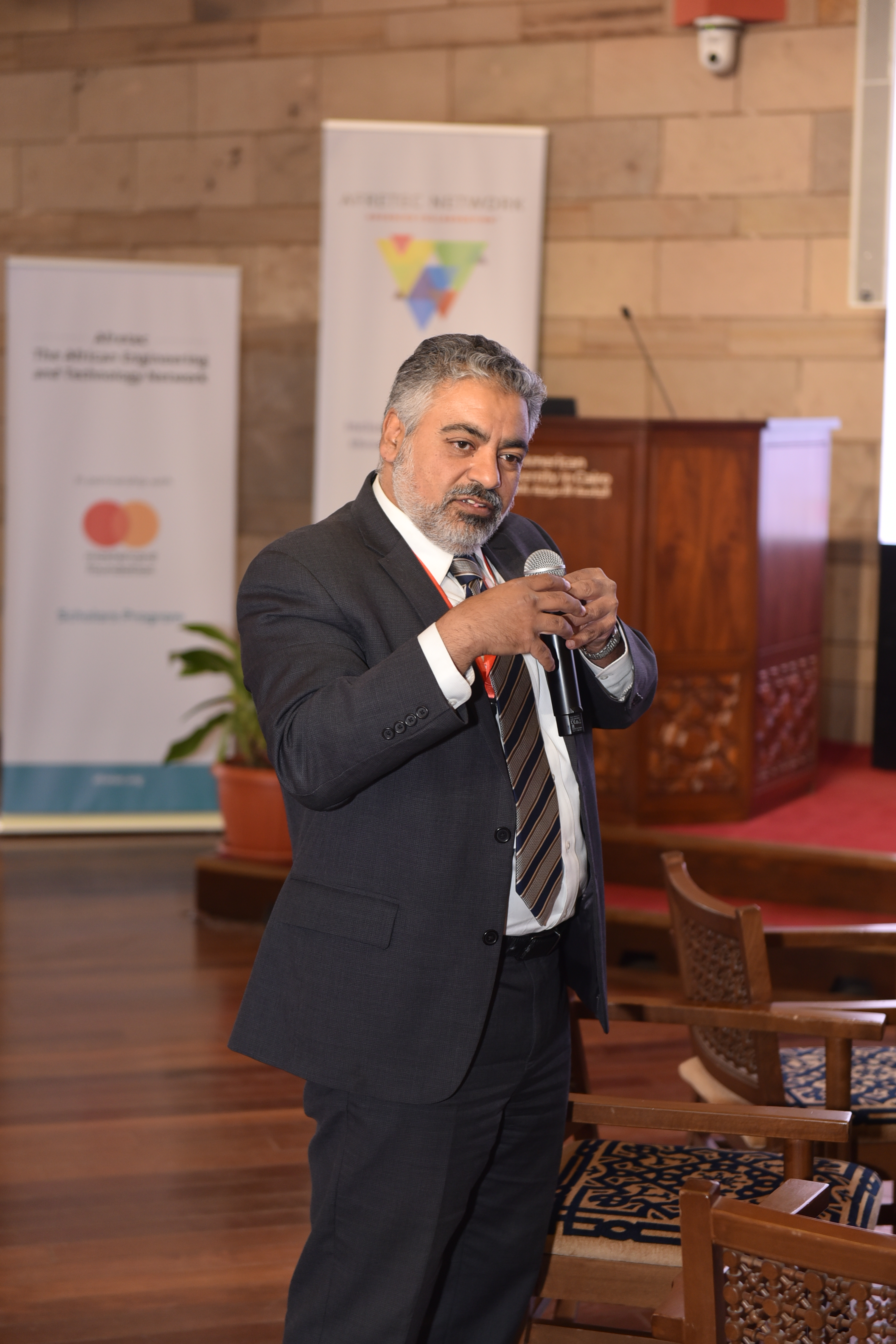 Professor Ismail stands and presents at the conference, speaking into a microphone and gesturing with his other hand.