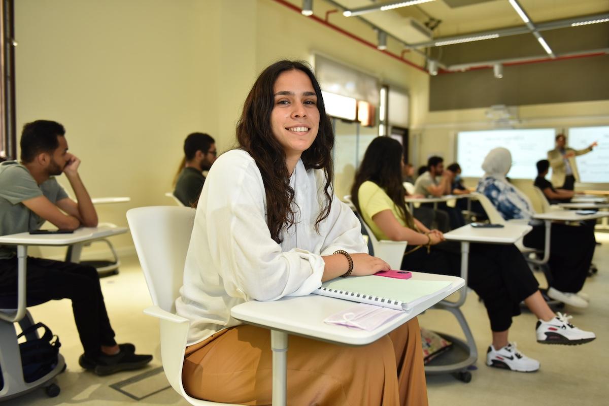 A student sits in a classroom with a notebook while listening to the instructor presenting at the front.