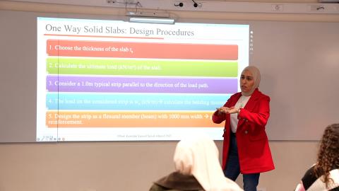 Woman in a red jacket and white hijab presents in front of a class