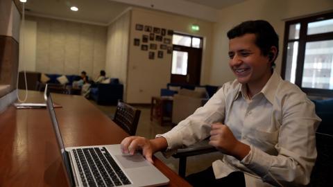 A male is sitting in a room and working on his laptop. He is smiling and he is wearing bluetooth earbuds