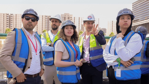 Five people, including students, in construction vests and helmets looking off into the distance