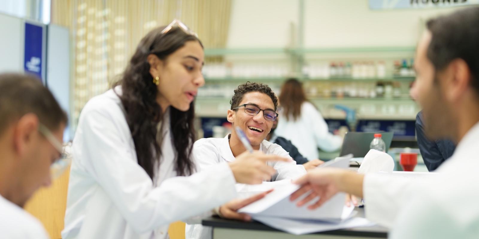 Students in white lab coats review papers together at a laboratory table, engaged in conversation.