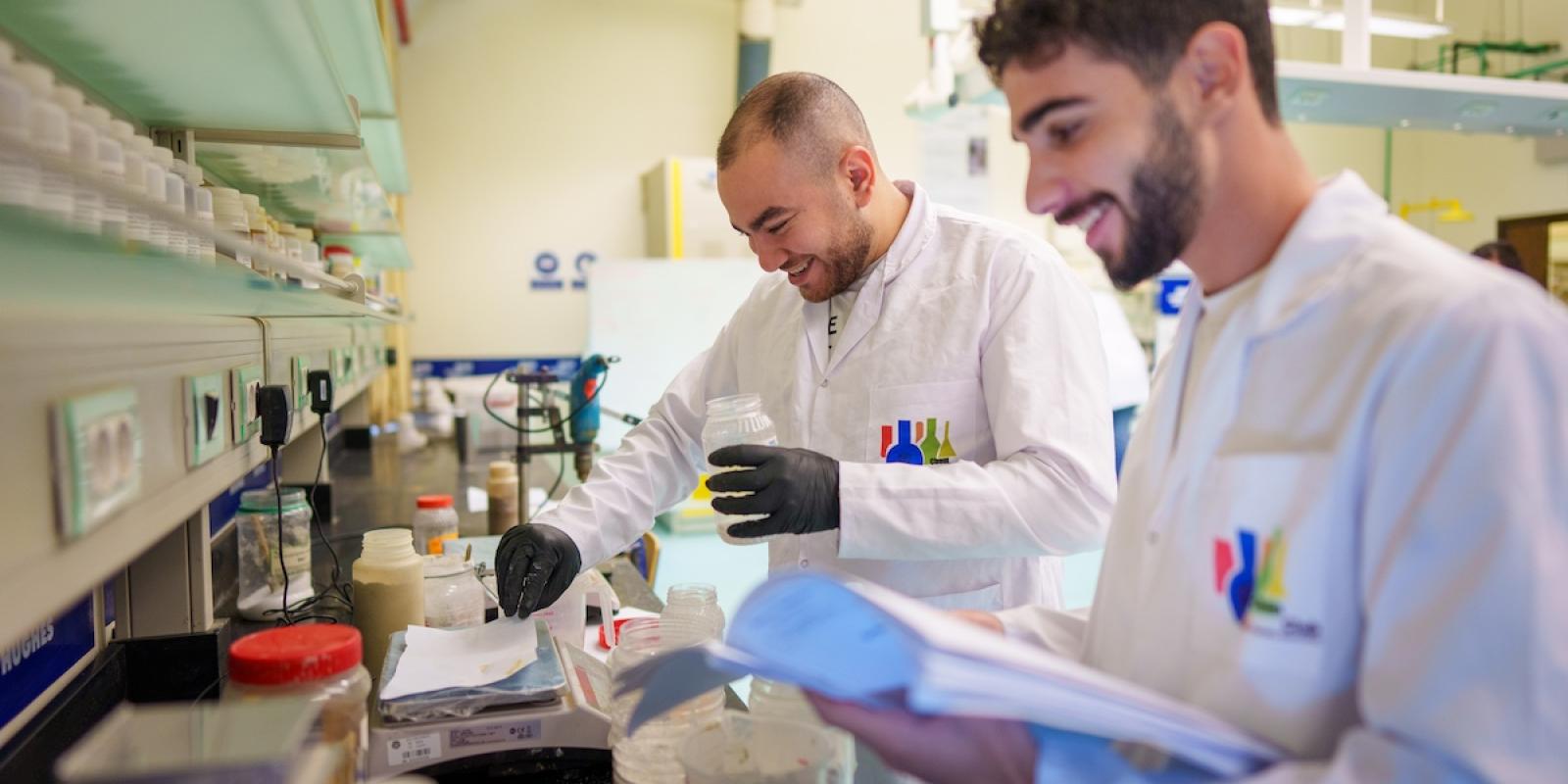 Two students wearing lab coats conduct an experiment in a laboratory while reviewing notes.