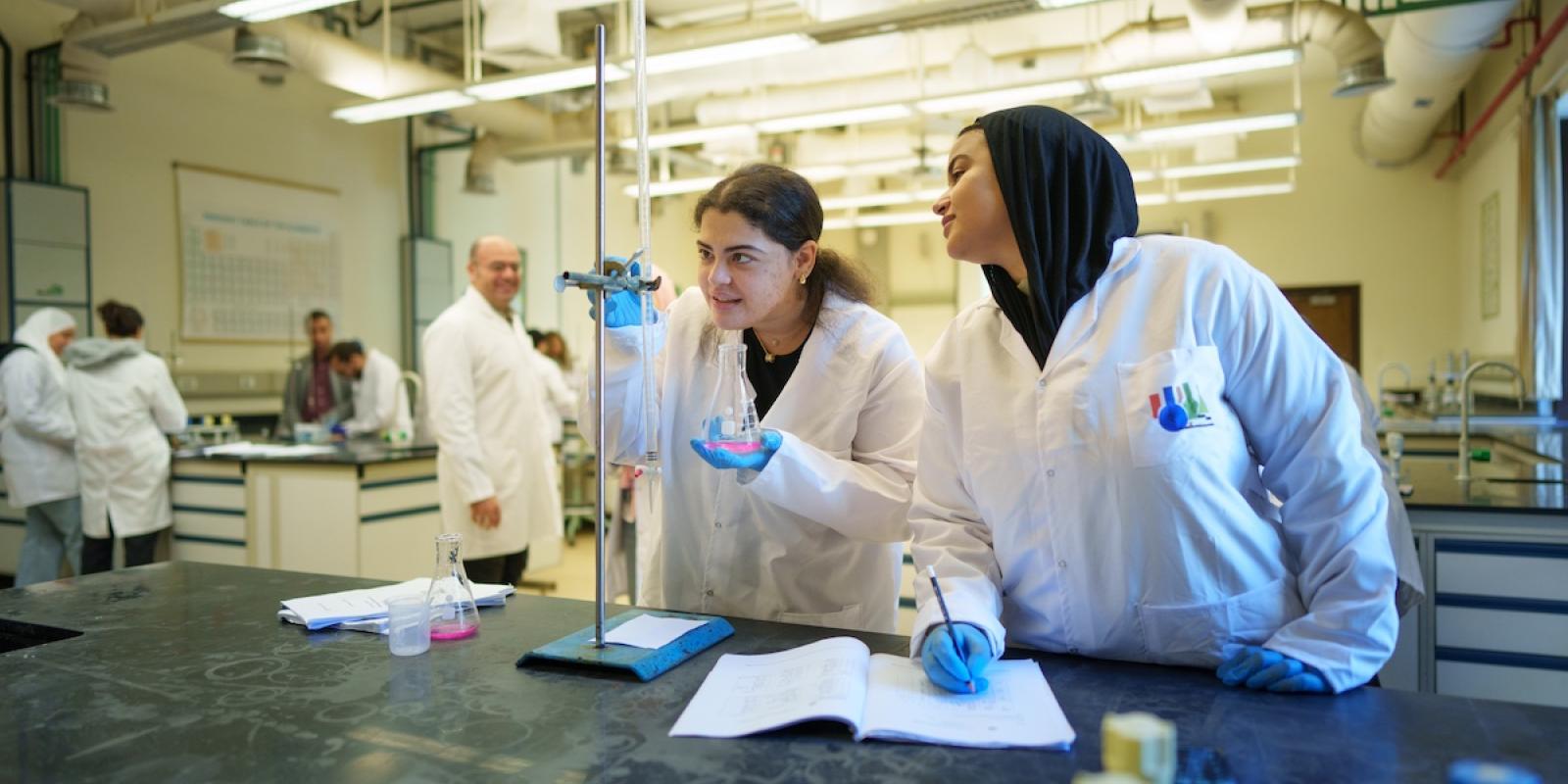 Two students in lab coats conduct a chemistry experiment using flasks and lab equipment while taking notes.