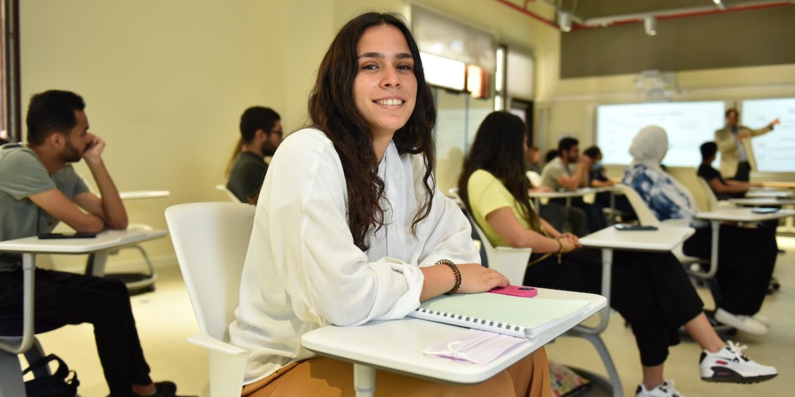 A student sits in a classroom with a notebook while listening to the instructor presenting at the front.