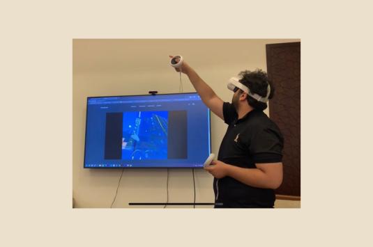 Man stands wearing a virtual reality headset and controllers while a screen behind him shows what he is seeing through the equipment