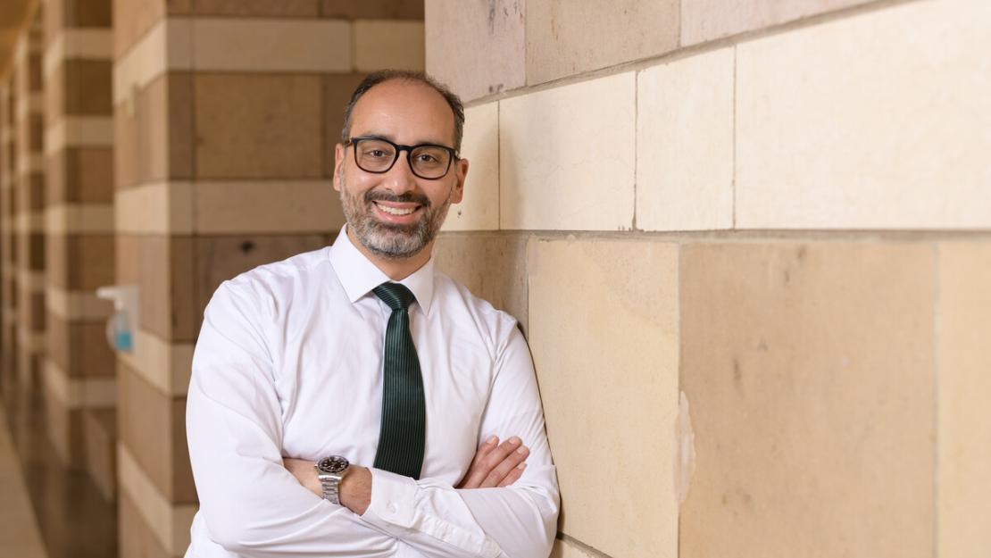 Smiling man in glasses wearing a button-up and blue tie leaning against a wall at AUC