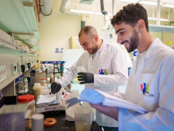Two students wearing lab coats conduct an experiment in a laboratory while reviewing notes.