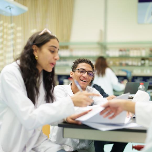 Students in white lab coats review papers together at a laboratory table, engaged in conversation.